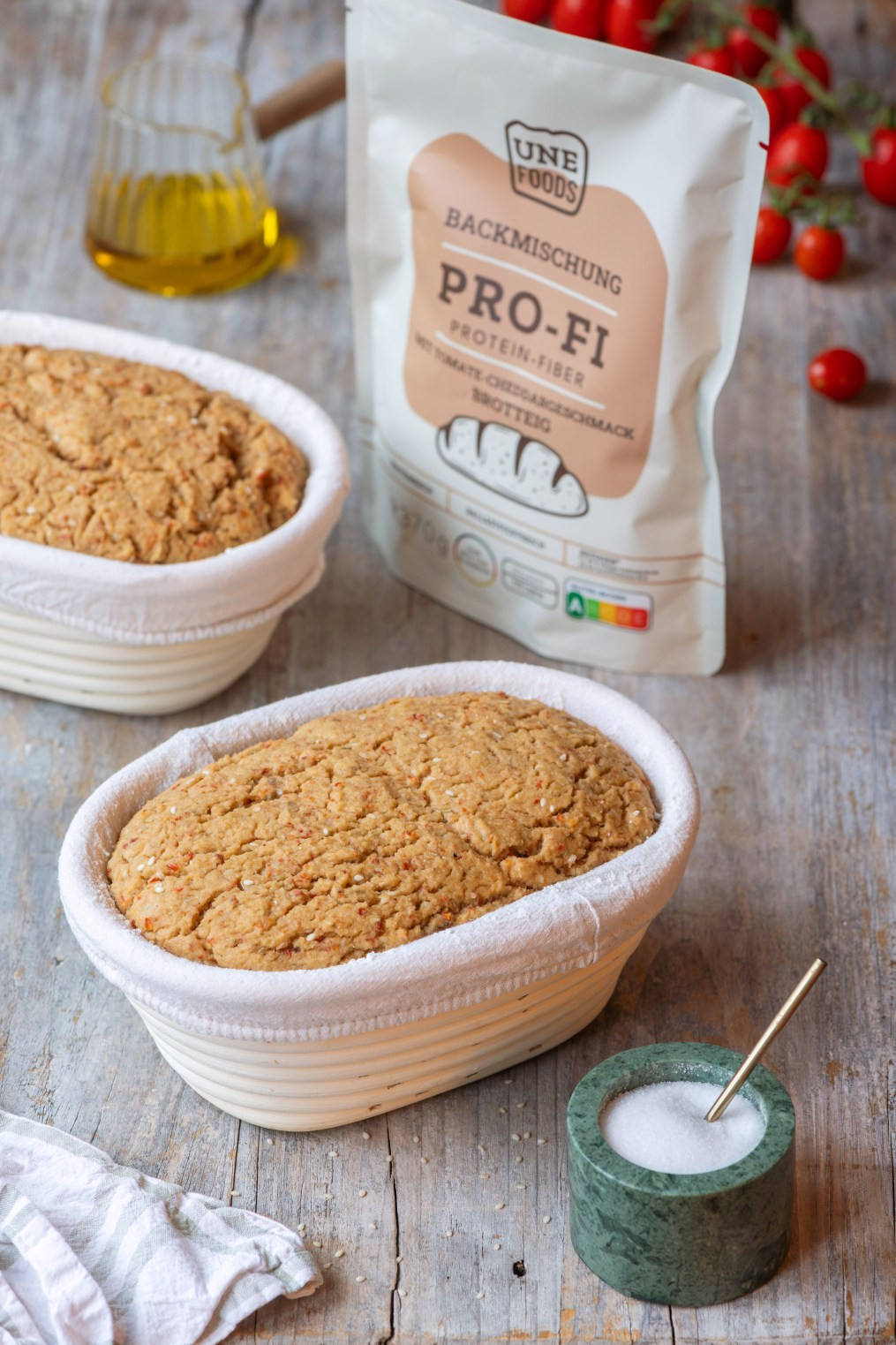 UNE Foods PRO-FI Protein-Fiber Brotbackmischung Tomate-Cheddar mit zwei gebackenen Broten in ovalen Gärkörbchen auf rustikalem Holztisch, dekoriert mit Olivenöl, Mehlschale und Kirschtomaten