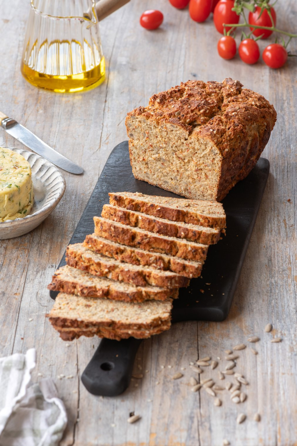 Aufgeschnittenes UNE Foods PRO-FI Tomate-Cheddar Proteinbrot auf schwarzem Schneidebrett mit Scheiben, dekoriert mit Olivenöl, Butter, Kirschtomaten und Sonnenblumenkernen auf Holztisch