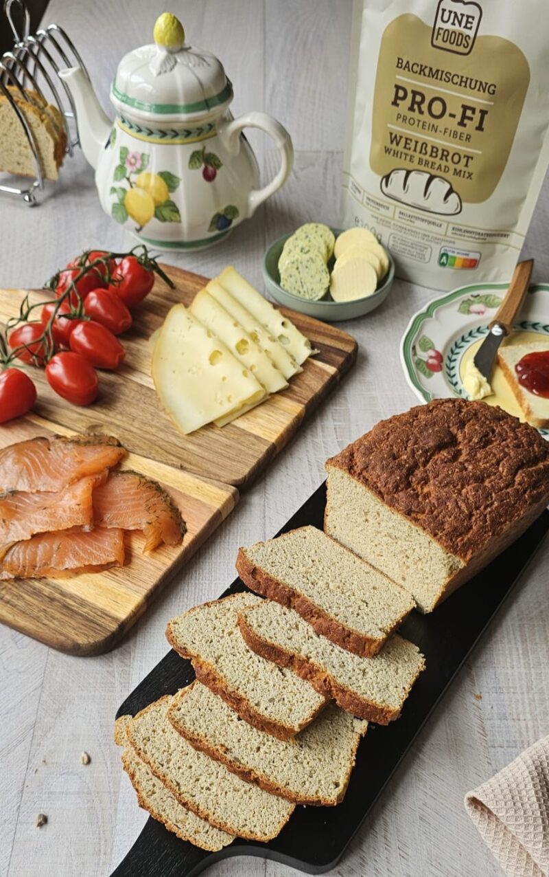 Frisch gebackenes und aufgeschnittenes Protein-Fiber Weißbrot auf hellem Holzbrett mit sichtbarer fluffiger Krume, im Hintergrund die PRO-FI Weißbrot-Backmischungsverpackung sowie eine kleine Schale mit Butter