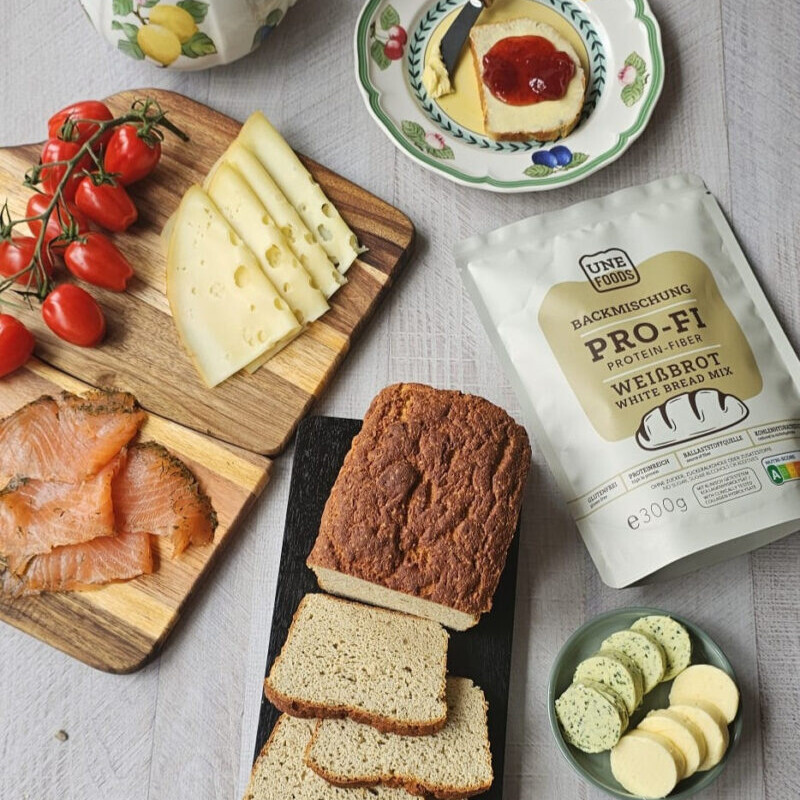 Frisch gebackenes und aufgeschnittenes Protein-Fiber Weißbrot auf hellem Holzbrett mit sichtbarer fluffiger Krume, im Hintergrund die PRO-FI Weißbrot-Backmischungsverpackung sowie eine kleine Schale mit Butter