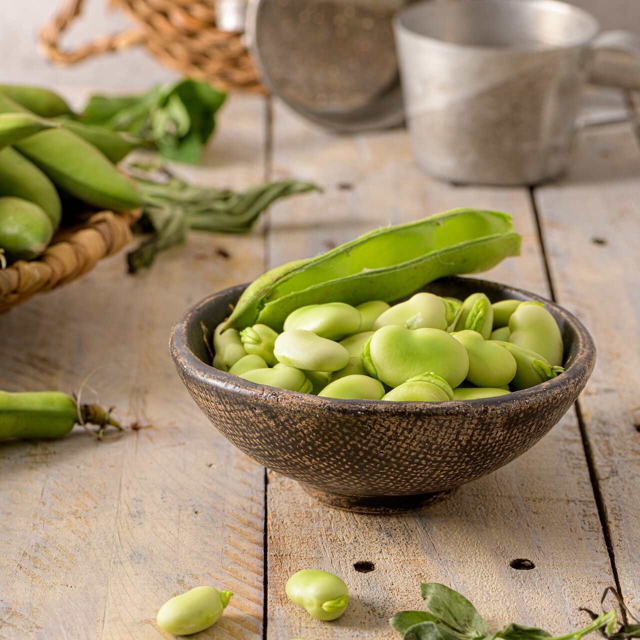 Frische gr&uuml;ne Ackerbohnen in rustikaler Holzschale auf hellem Holztisch, daneben Schere und frische Bohnenschoten, nat&uuml;rliche pflanzliche Proteinquelle f&uuml;r proteinreiche Backmischungen